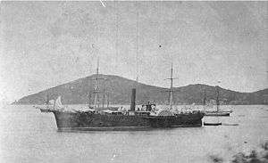  A black-and-white lithograph of the steamship USS De Soto, anchored in the harbor of Ponce, Puerto Rico. De Soto is in the foreground, facing left. De Soto is painted black or a very dark color, with the exception of her upper sidewheel guard which is white or very light in color. The ship has fine sweeping lines, a neatly rounded stern and slight clipper bow. She has two square-rigged masts with no sails, a tall, slim, raking smokestack amidships, just forward of the paddlewheels, and no apparent superstructure apart from what looks like temporarily rigged awnings on the main deck. The ship's walking beam engine can clearly be seen rising between the paddlewheels. In the distance are three sailing ships, one with a military appearance, and behind them two rounded, tree-covered hills which look almost uninhabited.