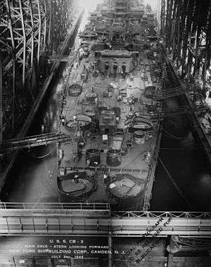 A large ship is being built. Seen from the stern, one of the 12-inch turrets equipped with three guns is visible. Most of the deck is cluttered with part of the ship in various stages of completion. Walkways connect the ship to the sides of the building ways.