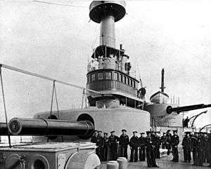 several men stand on deck next to a large turret, with a smaller one visible in the background