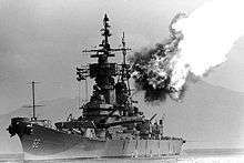 A black and white image; a large ship with gun barrels pointed to the right and up, flames and smoke can be seen emanating from the gun barrels.