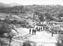 A large group of soldiers, some standing and some sitting, in a clearing below a hill covered in low grass and a few trees which can be seen in the background. In the middle ground a line of military vehicles are parked in a line on a road running front-on towards the camera, before hooking to the left in profile. Soldiers work around the vehicles, unloading stores.