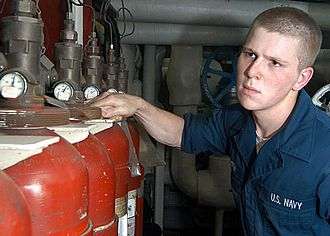 A sailor in coveralls wipes down red gas bottles