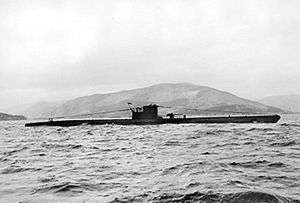 A side view of a surfaced submarine, seen in the middle distance; she is sailing in coastal waters and a mountainous landscape is in the background.