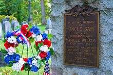 Close up of a grave stone (on the right) flanked by a red, white, and blue flower wreath (with ribbon and flag) on the left. Among other things, the stone reads "In Loving Memory of Uncle Sam"