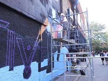Young people standing on a ladder spray painting art on the side of a building.