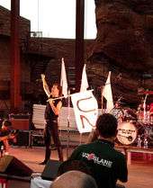 A man in black attire stands on a concert stage. With his left hand, he is holding a large white flag bearing the text "U2", while holding his right fist in the air. A red cliff is visible behind the stage.