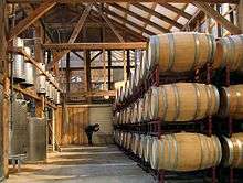 A wood-paneled warehouse with stainless steel fermentation tanks on the left, and rows of oak barrels on the right.
