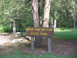 A brown wooden sign has Upper Pine Bottom State Park in yellow letters, with large tree trunks, rocks, and a picnic table in the background