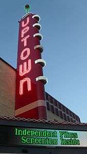 A red sign with the word "Uptown" illuminated in white neon. The sky is darkening. There is an electronic marquee that reads "Independent films screening inside".