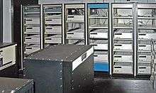 A room with a black box in the foreground and six control cabinets with space for five to six racks each. Most, but not all, of the cabinets are filled with white boxes.