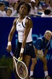 A black woman is serving the ball, and is wearing a white sleeveless top and blue skirt