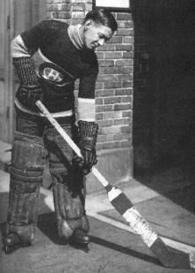 Profile of a goaltender in full uniform looking down at his stick. He is wearing thick pads around his legs, and padded gloves that reach near his elbows.
