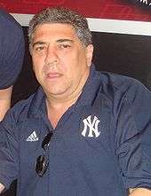 A man with graying hair wearing a navy blue shirt which is holding his sunglasses. Logos for Adidas and the New York Yankees appear on his shirt