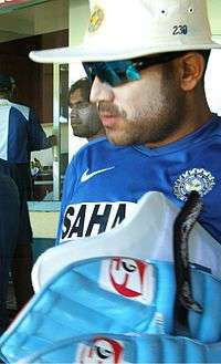 A man in the blue Indian cricket practice kit, wearing sun cream, sunglasses and a hat carrying his batting pads. Others can be seen in the background.