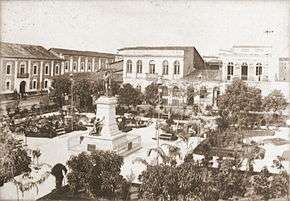 A photograph overlooking a large city plaza containing low trees and walkways in the center of which stands a sculpture on a high plinth