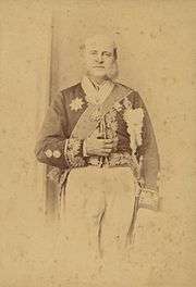 Photograph of an older man with long sideburns standing in a heavily embroidered cutaway jacket adorned with various medals and wearing a sash of office