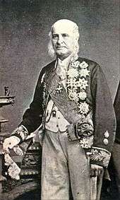 A tintype photograph showing an older, white-haired man standing in formal dress with a sash of office and a cutaway coat with embroidered sleeves which is covered in medals
