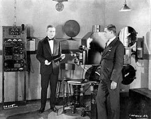 Two suited men stand in a studio with a large film projector and other electrical equipment. The man on the left is holding a large phonograph record.