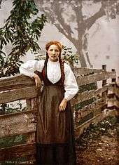 A girl with auburn hair in pigtails poses next to a rude wooden fence. She wears a white ruffled shirt and a long brown dress.