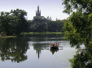 A small row boat navigates a wide river, while a forest stands in the background, hiding a large tower.