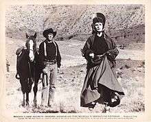 Printer poster with a photograph and text underneath. A man is standing holding the reins of his horse. A woman is running towards the camera. They are in a dry landscape with a stony canyon wall in the background. The text consists of a copyright notice and a license for periodicals to reproduce the photograph.