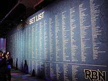 A long list of songs and artists against a blue background on a wall. About three quarters of the way down the wall is a white line; the songs above the line are labeled "Rock Band Setlist" while the songs below it are labeled "RBN".