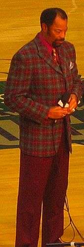 A man, wearing red and green jacket with a red shirt and tie, is standing on a basketball court while holding a microphone.