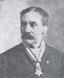 Head and shoulders of a white man with parted hair and a large mustache, wearing a star-shaped medal from a ribbon around his neck.