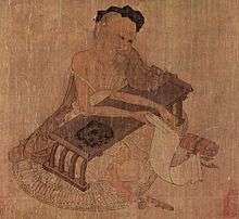 Elderly Chinese man, seated at a low writing desk and holding a scroll