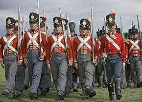 Photo of men dressed in British uniforms of the early 1800s. They wear red coats with white cross belts, gray trousers, and black shakos.