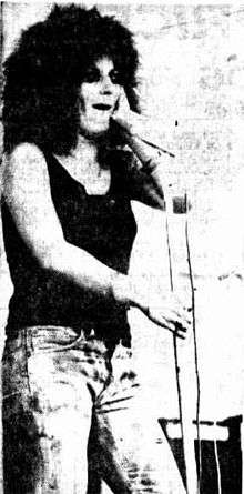 A 20-year-old woman is singing at a microphone and staring into the distance. She wears a dark singlet-top and jeans and has her hair in an afro. Her right arm is slightly bent as she holds the microphone stand. Her left hand is up near her left cheek.
