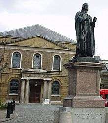A bronze statue of John Wesley dressed in robes and preaching bands in the foreground, with a Georgian chapel in the background