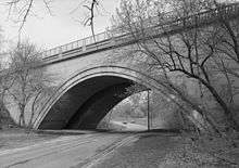 Looking slightly up at a black and white image of a Neoclassical concrete bridge over a road