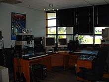 A musician's office with a solid wall and a wall containing windows shown, a microphone stand, an electric guitar, a mixing board, a keyboard, two computer monitors and a computer, a poster of the Transformers franchise