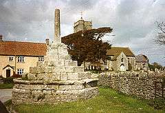 Stone steps up to a stone shaft which would once have had a cross at the top. To the left are yellow painted houses. To the right is an old stone church with a square tower partially obscured by trees.