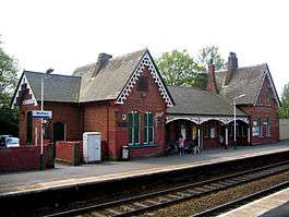 Two sets of rails between platforms beyond which is a brick building with two gables and a canopied area between them; a sign to the left says "Widnes"