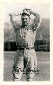 A man wearing a lightly colored baseball uniform holds his hands above his head, winding up to throw.
