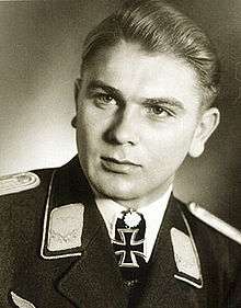 The head and shoulders of a young man, shown in semi-profile. He wears a military uniform with an Iron Cross displayed at the front of his white shirt collar. His hair is blond, short and combed to his back, his nose is long and straight, and his facial expression is emotionless; looking to the right of the camera.