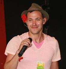 A man in a pink t-shirt smiling and holding a microphone.