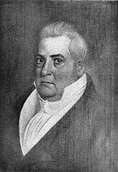 A slightly portly, clean-shaven man with long gray hair wearing a high-collared, white shirt and black jacket