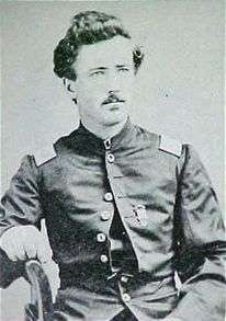 Head and torso of a white man sitting sideways in a chair, his right arm resting on the chairback. He has a thin mustache, curly hair, and is wearing a military jacket.