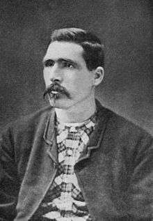 Head and shoulders of a dark-haired white man with a thin, drooping mustache, wearing an unbuttoned dark jacket over a checkered shirt.