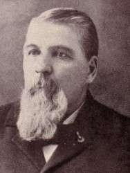 A white man with a full, light beard and dark suit, looking to the left.