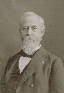 Head and shoulders of an older white man with a bushy beard wearing a suit coat, vest, and bow tie.