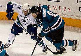 An ice hockey player dressed in a white and blue jersey attempting to impede another player dressed in a teal and black jersey with his stick.