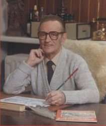 Man with short hair and horn-rimmed glass, and wearing a cardigan sweater and tie, sits at a desk holding a pencil and drawing.