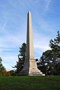 Light colored stone obelisk that stands on a wide base, on which is inscribed family names.