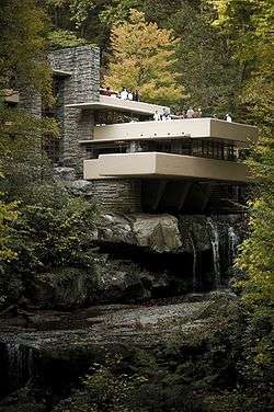 A modernist-style house sits nestled in the woods, with a multilevel terrace hanging over a waterfall