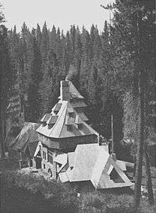A monochrome photograph of a 75-foot-tall building with castle-like features, seen from a nearby higher elevation, surrounded by tall pine trees.