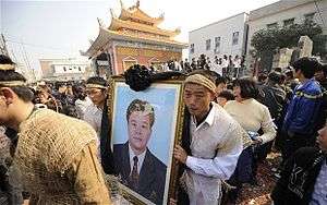 Procession led by some Asian people dressed in white/hemp carrying a photograph of a man in a suit.